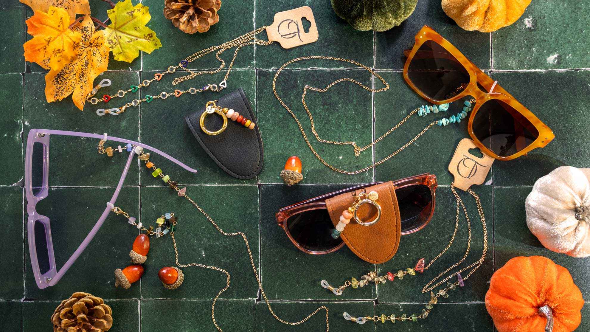 Feestelijke headerfoto met kleurrijke OT brillenkoordjes, brillen-houders, en zonnebrillen, omringd door herfstdecoraties zoals bladeren, eikels, en pompoenen.