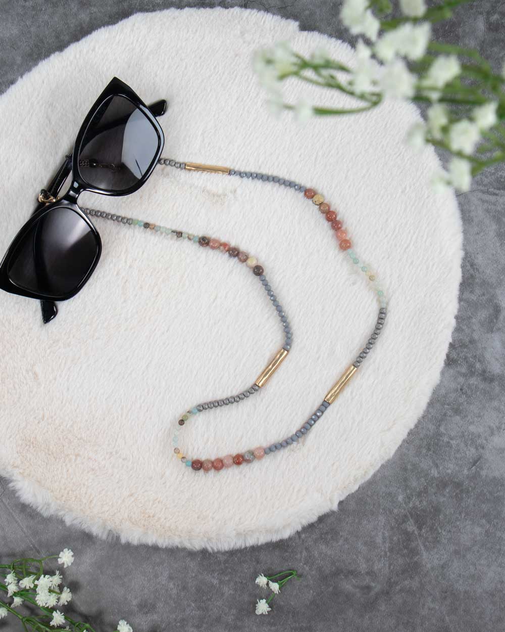 Een brillenkoord met pastel kralen op een wit kleedje en als decoratie een zonnebril.